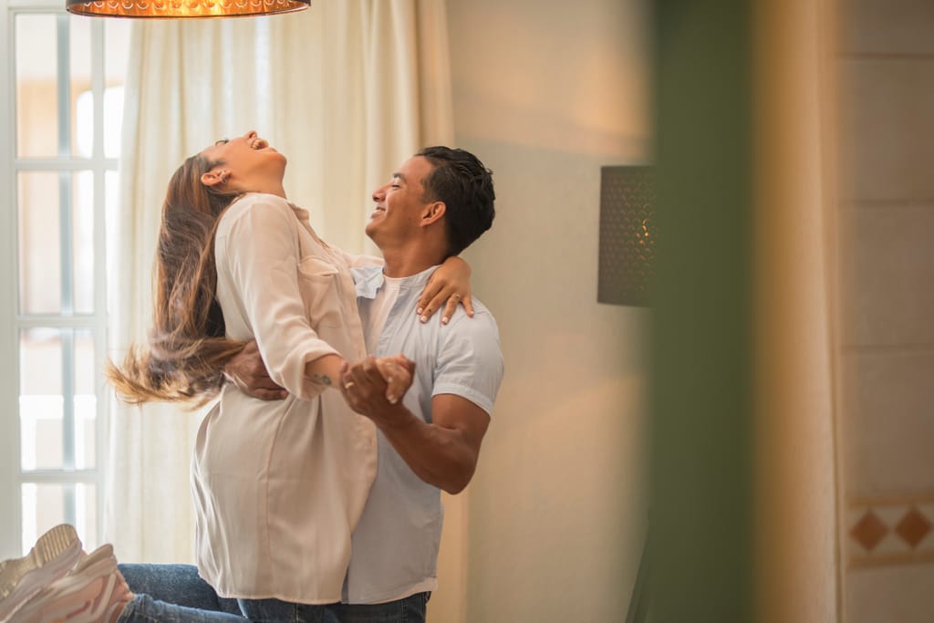 Pari tanssii olohuoneessa; mies nostaa naista ilmaan ja molemmat nauravat, valoisan kodin ja verhojen luodessa lämpimän tunnelman.