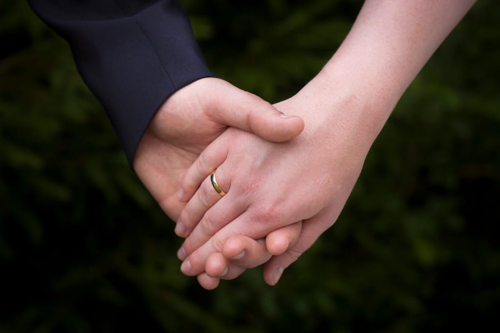 Kaksi avioliitossa olevaa aikuista pitää toisiaan kädestä ulkona; etualalla näkyy sormus toisen kädessä, taustalla vihreä, epätarkka luonto.