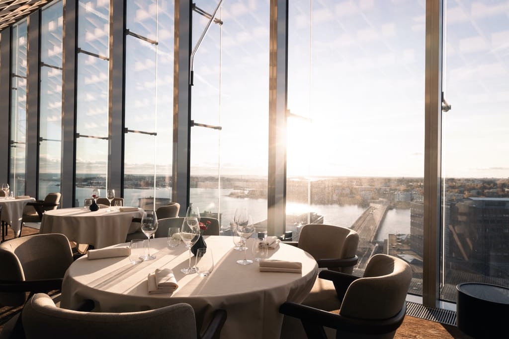 Ravintolasali, jossa on isot lasiset ikkunat. Pyöräeä pöytä jossa on valkoinen pöytäliina ja kattaus.