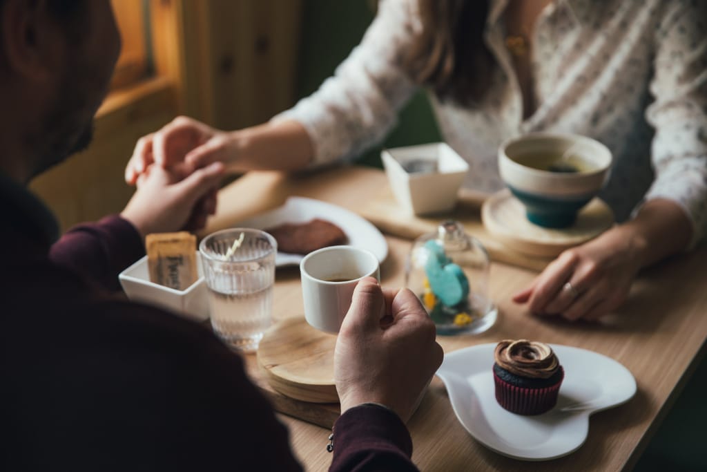 Lähikuva kahvilapöydästä, jossa kaksi ihmistä istuu vastakkain. He pitävät toisiaan kädestä pöydän yli. Pöydällä on kahvikuppeja, leivoksia ja pieniä astioita, mikä viittaa rennolle ensitreffitilanteelle.
