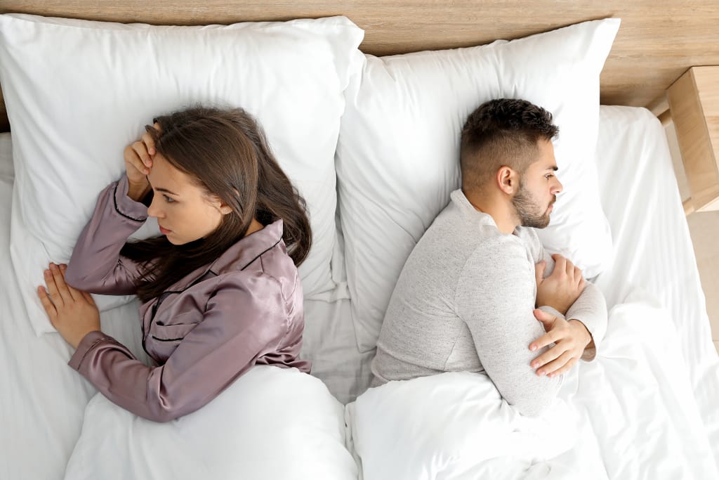 Pariskunta makaa sängyssä selät vastakkain; nainen näyttää huolestuneelta ja mies pitää käsiään ristissä, mikä viittaa parisuhteen jännitteisiin.