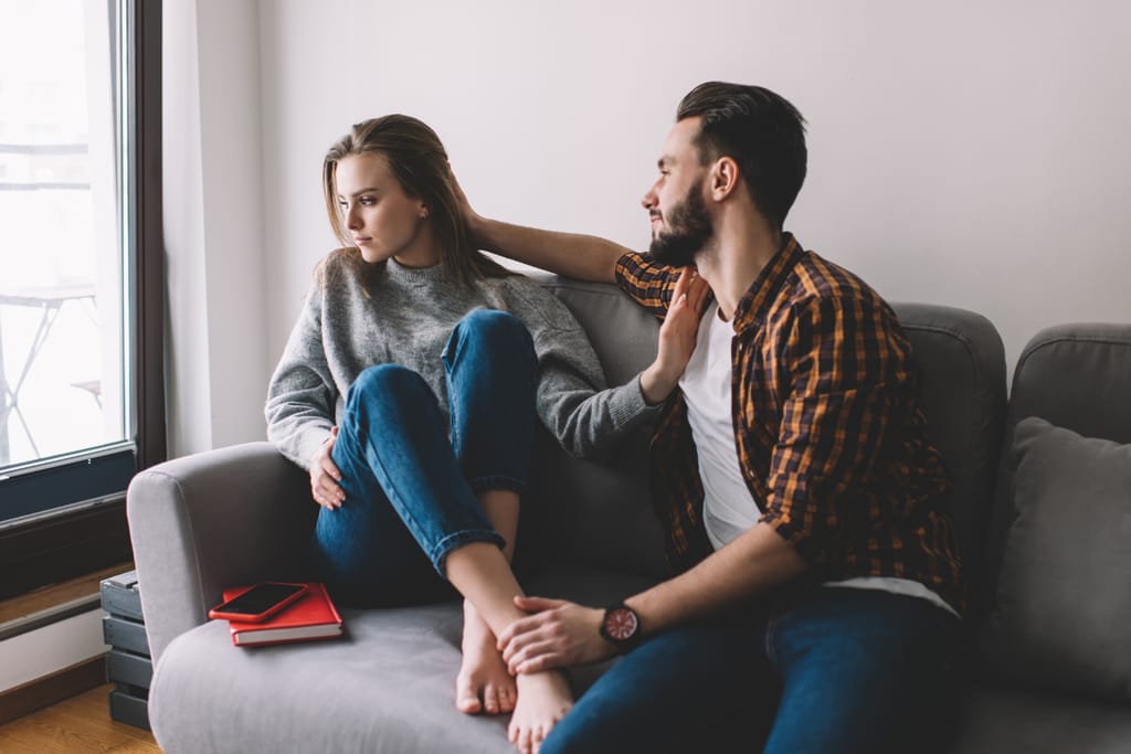 Nuori nainen istuu sohvalla polvet koukussa ja katsoo poispäin, kun vieressä istuva mies koskettaa häntä rauhoittelevasti käsivarteen ja jalkaan; naisen kehonkieli on sulkeutunut ja etäinen.