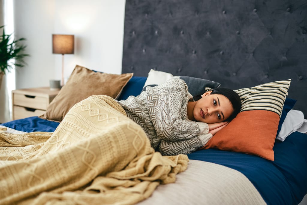 Nainen makaa yksin sängyssä peiton alla kyljellään ja katsoo ajatuksiinsa vaipuneena eteenpäin makuuhuoneessa.