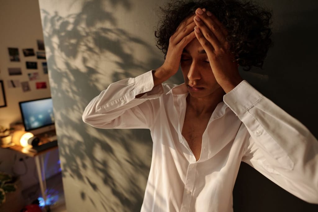 Hoikkavartaloinen mies pitelee päätään käsiensä välissä kuin yrittäisi koota itseään, ja hänen valkoisen kauluspaitansa ylimmät napit ovat auki. Tausta on asunto, jonka kulman takana on tietokoneen näyttö päällä ja seinällä valokuvia tai tauluja. Iltavalaistus.