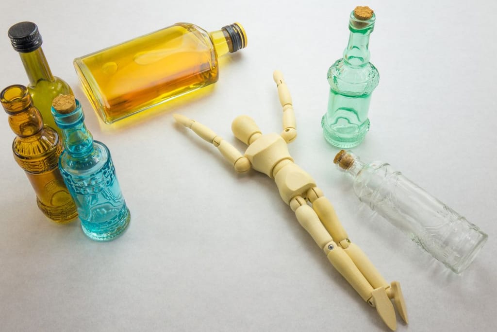 Puinen ihmishahmonukke makaa selällään lasipullojen keskellä. Asetelma viittaa humalatilaan.