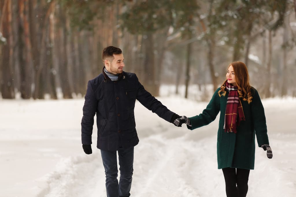 Pariskunta kävelee käsi kädessä lumisessa metsämaisemassa talvivaatteisiin pukeutuneina. He katsovat toisiaan hymyillen, ja tunnelma on rauhallinen, romanttinen ja yhteenkuuluvuutta korostava.