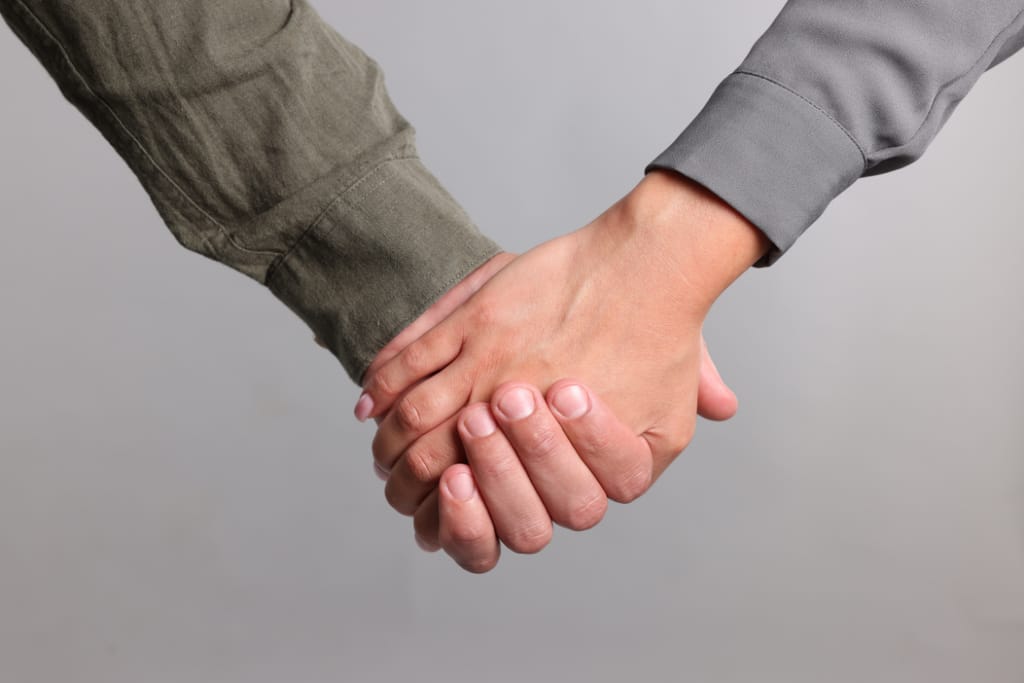 Kaksi henkilöä pitää toisiaan kädestä vaaleaa taustaa vasten, lähikuva käsistä ja hihoista.