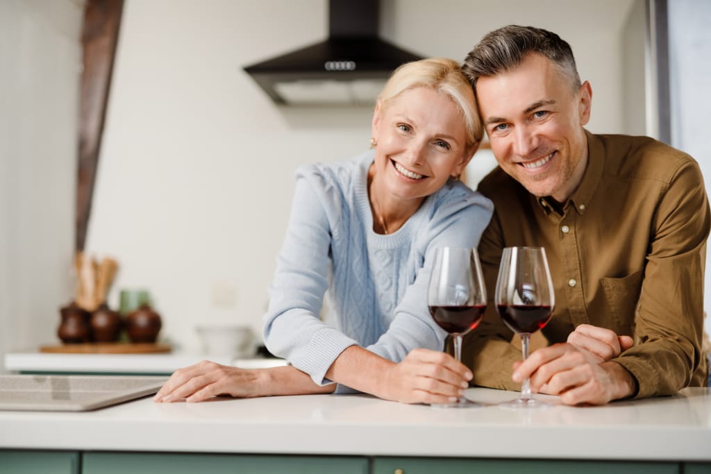 Kaksi aikuista hymyilevää henkilöä nojaa keittiön tasoon ja pitelee viinilaseja, katsoen kameraan valoisassa kodissa.
