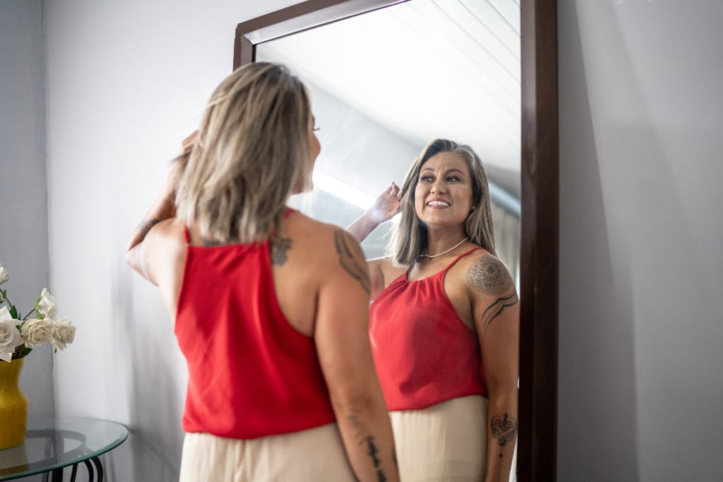 Nainen seisoo peilin edessä ja hymyilee omalle peilikuvalleen. Hänellä on punainen hihaton paita ja vaalea hame, ja hänen käsivarsissaan näkyy tatuointeja. Nainen kohottaa kättään hiustensa luo ja katsoo itseään itsevarmasti valoisassa huoneessa.