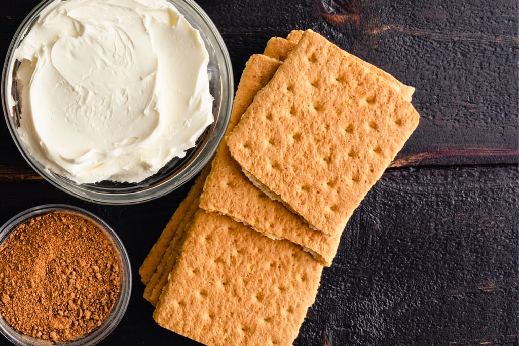 Tummalla alustalla on kulho kreikkalaista jogurttia, vieressä murennettua Biscoff-keksimaustetta sekä pino suorakaiteen muotoisia Biscoff-keksejä.