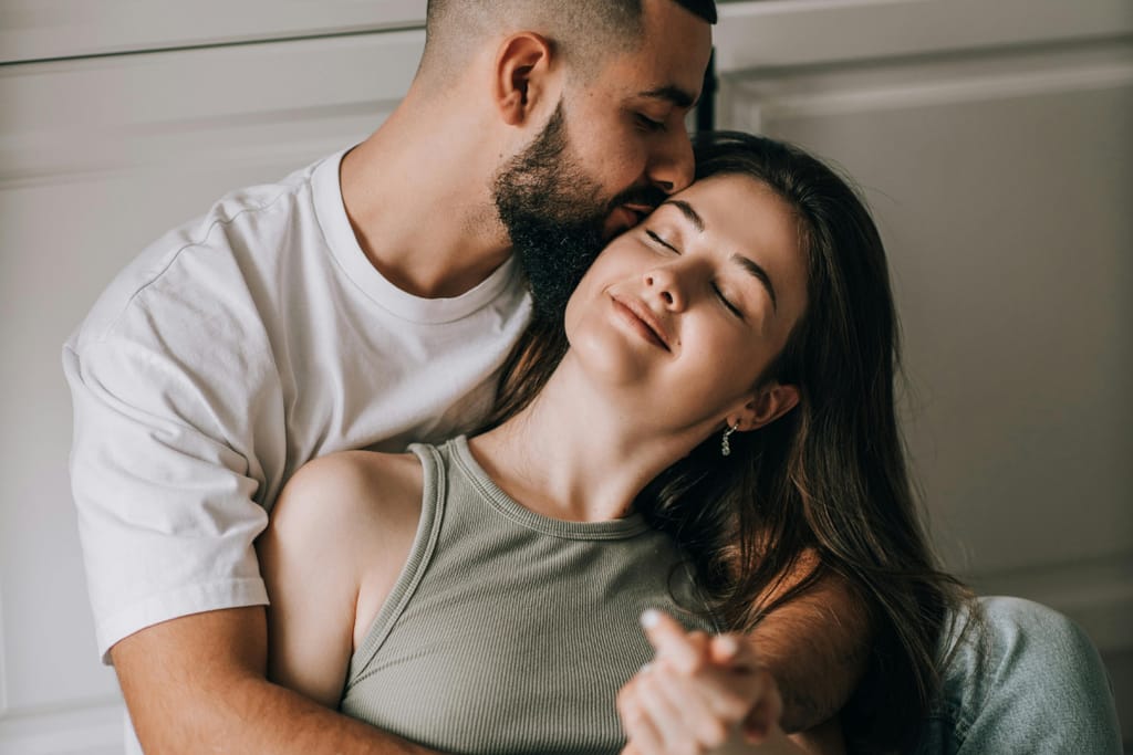 Mies pitää naista sylissään ja suutelee tätä hellästi ohimolle, nainen nojaa taaksepäin silmät kiinni ja hymyilee rauhallisesti, hetki kuvastaa turvallisuutta, kiintymystä ja emotionaalista läheisyyttä.