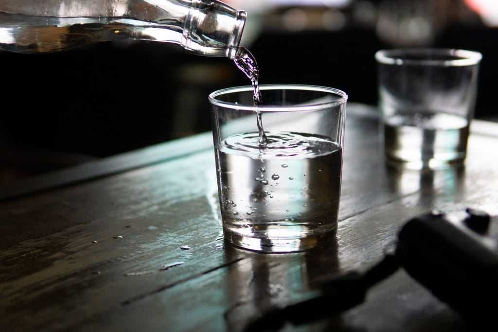 Kirkasta nestettä kaadetaan pullosta lasiin hämärässä ympäristössä. Kuvassa näkyy kaksi juomalasia pöydällä, ja asetelma viittaa alkoholiin ja juomiseen.