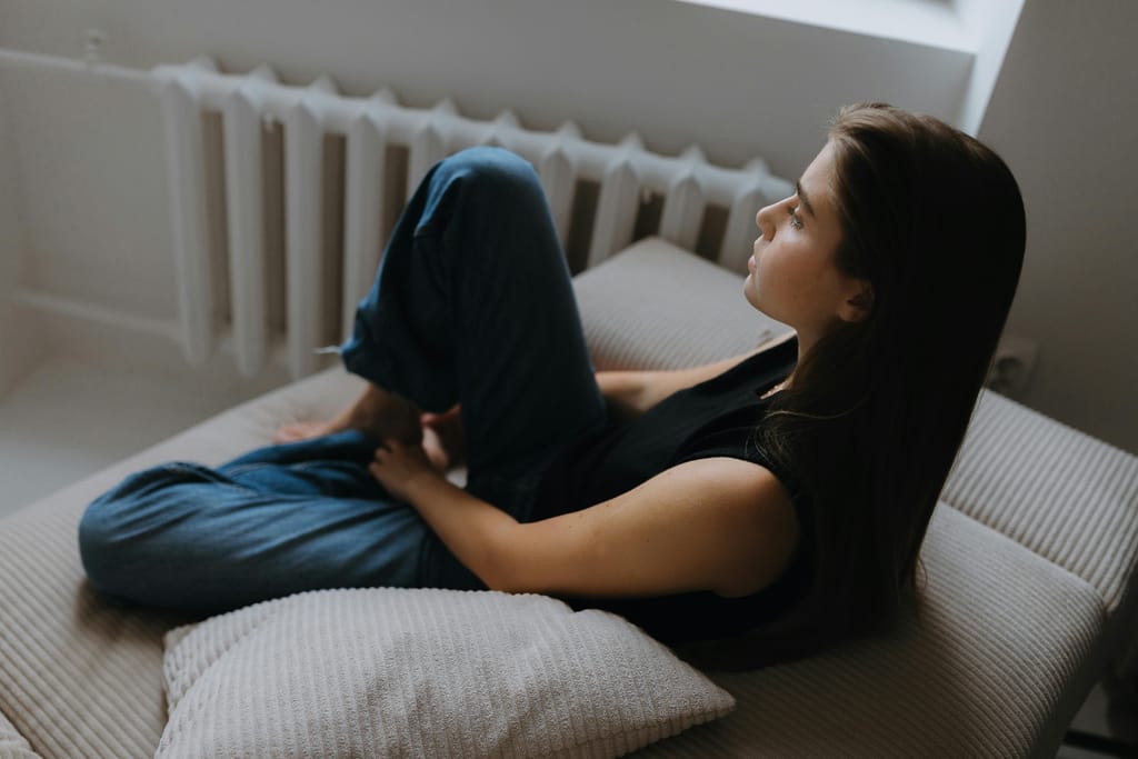 Nuori nainen istuu sängyllä polvet koukussa ja katsoo sivulle ikkunan suuntaan; huone on hämärä ja rauhallinen, ja asento viestii yksinäisyydestä ja mietteliäästä mielialasta.