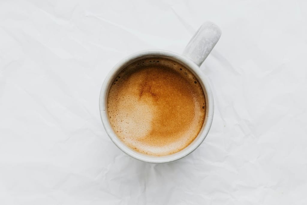 Kupillinen kahvia kevyesti vaahdotettuna vaalealla taustalla. Kuppi on saman sävyinen kuin pöytä. Kuva on otettu yläviistosta.