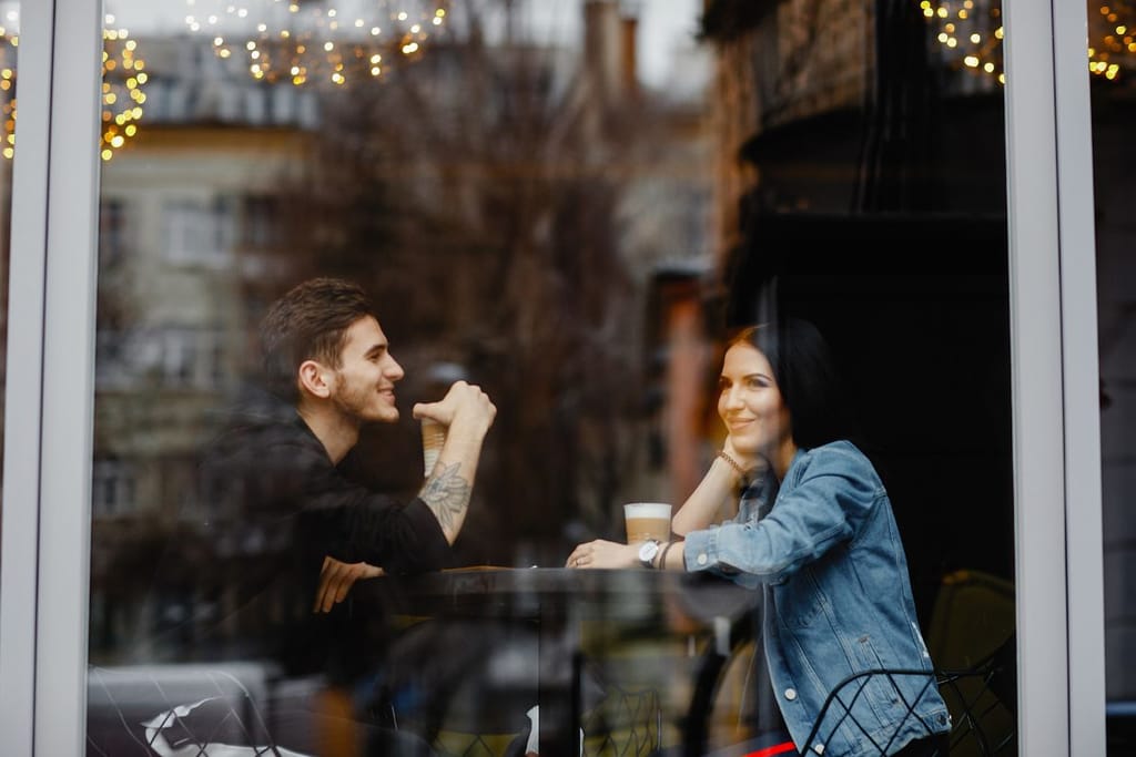 Nainen ja mies istuvat kahvilan ikkunan viereisessä pöydässä. Nainen näyttää vaivaantuneelta ja katselee ulos ikkunasta, kun mies selittää jotain itsetyytyväisenä.