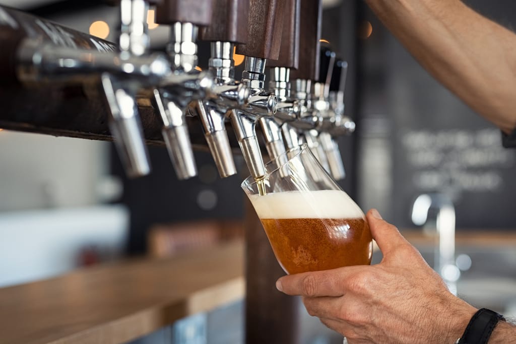 Lähikuva käsistä kaatamassa vaaleaa hanaolutta lasiin, jossa muodostuu vaahtokruunu; taustalla näkyy useita metallisia oluthanoja baaritiskillä.