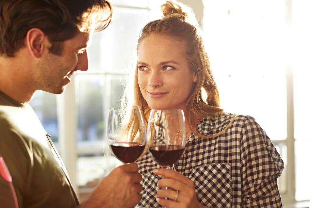 Mies ja nainen seisovat vastakkain valoisassa huoneessa ja kilistävät punaviinilaseja katsoen toisiaan hymyillen.