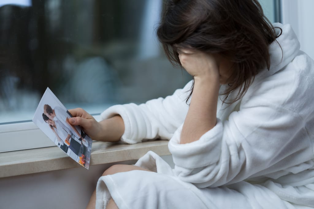 Ikkunan äärellä istuva henkilö pitää kädessään valokuvaa ja peittää kasvonsa toisella kädellä, ilme surullinen ja mietteliäs.