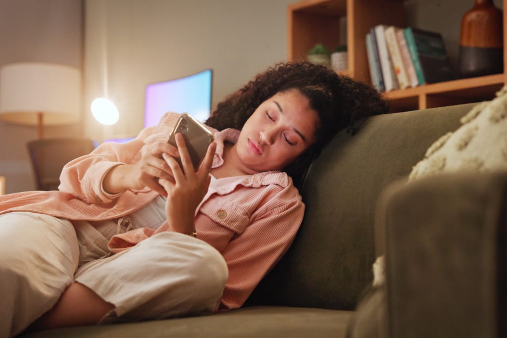 Nuori nainen makaa sohvalla olohuoneessa ja selaa puhelintaan silmät väsyneinä; ympärillä on lämmin valaistus, kirjahylly ja tietokonepöytä.