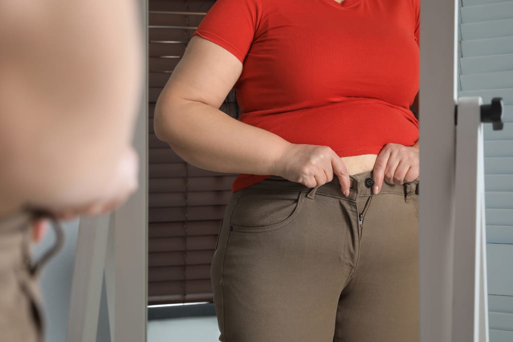 Henkilö seisoo peilin edessä ja yrittää sulkea liian tiukkoja housuja punainen paita päällä.