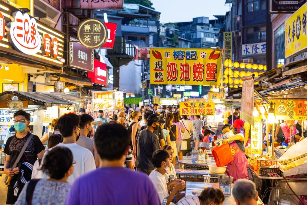 Vilkas aasialainen yömarkkina täynnä ihmisiä. Kapeaa katua reunustavat kirkkaasti valaistut ruokakojut, joissa myydään katukeittiön herkkuja. Kyltit ovat kiinankielisiä ja värikkäitä, ja taustalla näkyy tiiviisti rakennettuja taloja. Ihmiset jonottavat, syövät ja kulkevat kojujen välissä iltahämärässä.