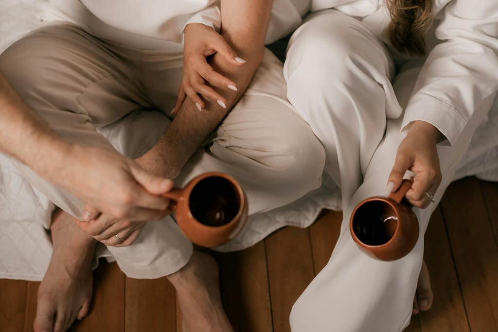 Pariskunta valkoisissa kylpytakeissa istuu sohvalla suuren ikkunan äärellä, hymyilee ja katsoo ulos samalla kun he pitelevät kahvikuppeja luonnonvalossa.