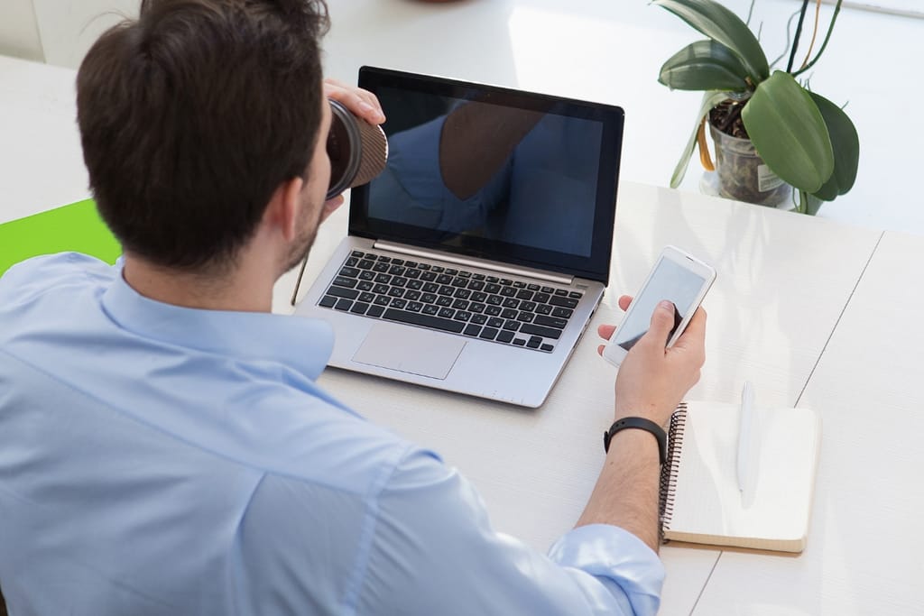 Ylhäältä kuvattu näkymä miehestä työpöydän ääressä, joka juo kahvia, katsoo älypuhelinta ja käyttää kannettavaa tietokonetta muistikirjan vieressä; tilanne symboloi etätyötä, moniajoa ja LinkedIn treffailu -tyyppistä ammatillisen ja henkilökohtaisen elämän sekoittumista.