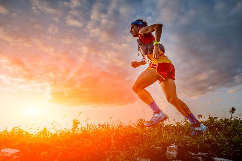 Ihminen maastojuoksee niittyisessä maastossa auringon laskussa. Hänellä on pääpanta, painovyö, urheilukello ja säänmukainen lenkkeilyvarustus. Taivaalla on pilviä.