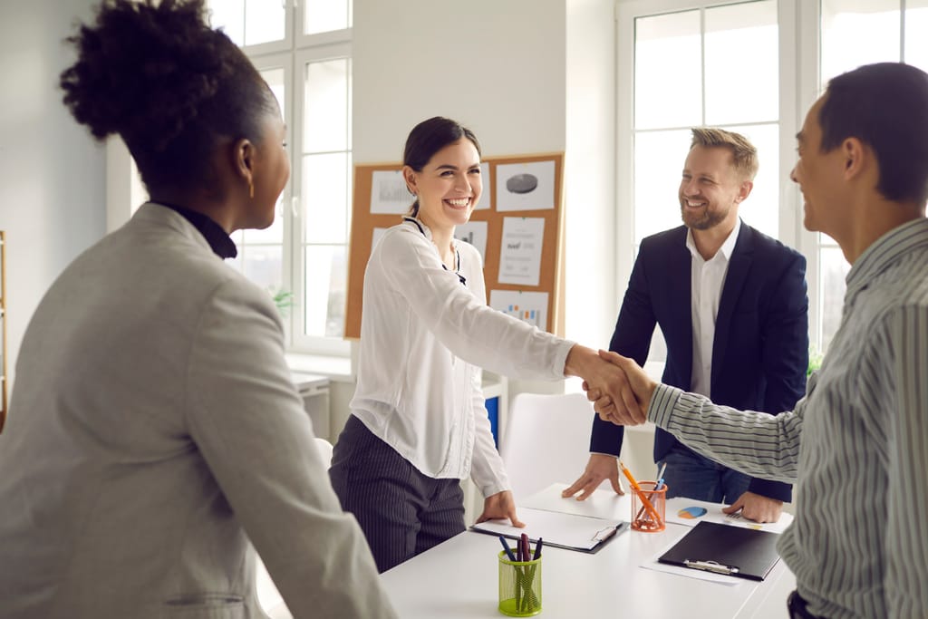 Neljän hengen työryhmä seisoo valoisassa toimistossa pöydän äärellä; keskellä oleva nainen kättelee miestä, ja muut tiimin jäsenet seuraavat tilannetta hymyillen, pöydällä näkyy asiakirjoja ja toimistotarvikkeita.