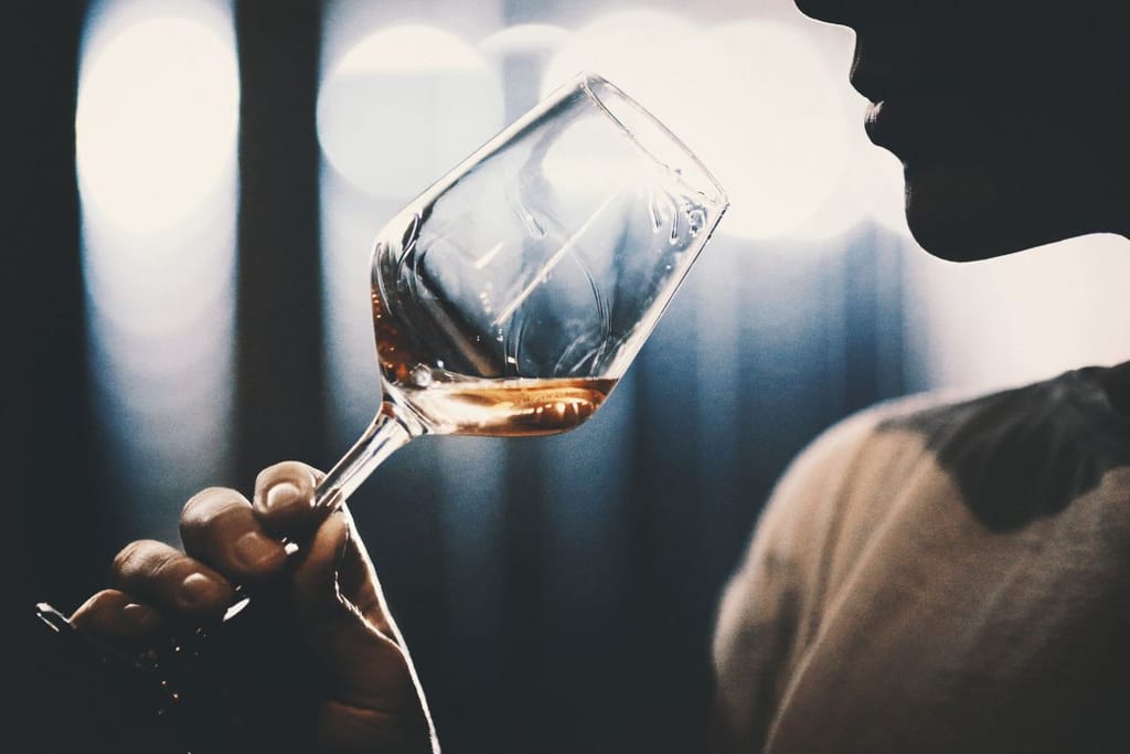Henkilö maistaa viiniä viinilasista hämärässä valaistuksessa, kasvot sivuprofiilissa ja taustalla epäteräviä valopisteitä.