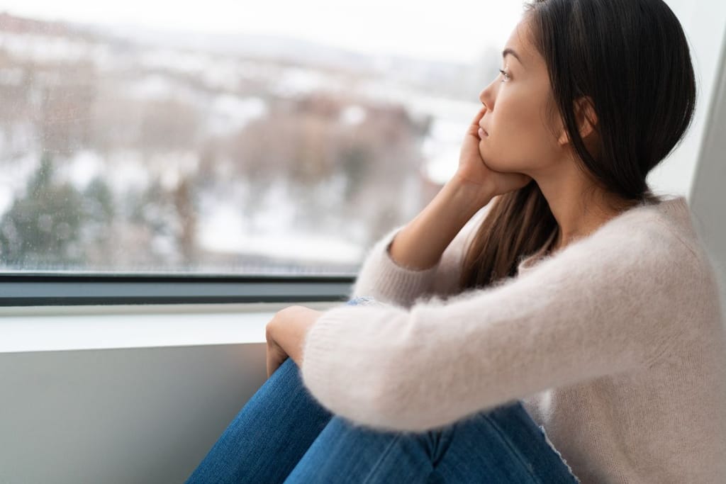 Nainen istuu ikkunan vierellä ja nojaa polviinsa ja leuka lepää hänen oikean kätensä alla. Nainen katselee talvista, urbaania maisemaa. 