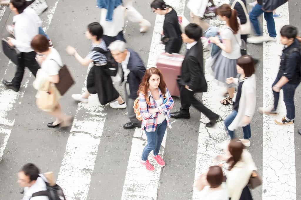 Nainen seisoo suojatiellä vilkkaassa kaupunkiympäristössä muiden ihmisten kulkiessa ohi.