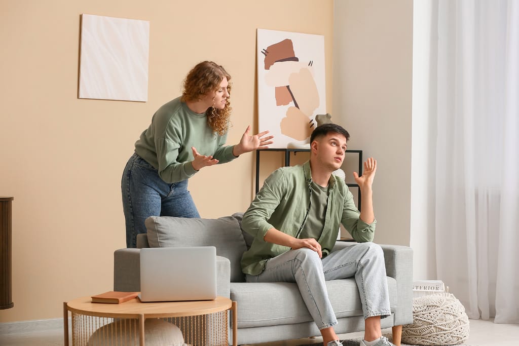 Nuori nainen seisoo olohuoneessa ja elehtii turhautuneena miehelle, joka istuu sohvalla ja näyttää välinpitämättömältä. Mies nojaa taaksepäin ja kohottaa kättään puolustautuvasti, kun nainen yrittää puhua. Tilanne kuvastaa ristiriitaa ja vallankäyttöä läheisessä ihmissuhteessa.