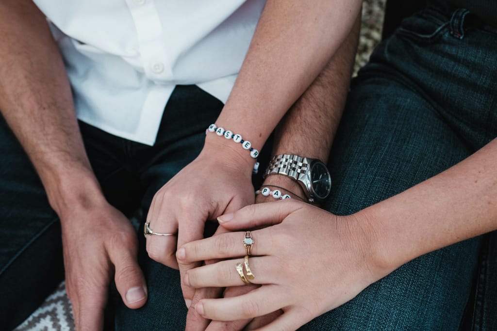 Lähikuva pariskunnan käsistä, jotka lepäävät toistensa päällä. Molemmilla on sormuksia ja rannekoruja, ja käsien kosketus viestii läheisyydestä ja sitoutumisesta.