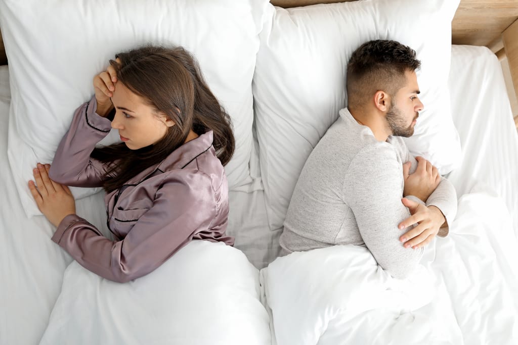 Nuori pari makaa sängyssä selät vastakkain. Nainen katsoo sivulle käsi otsallaan ja mies on kääntyneenä poispäin kädet ristissä, molemmat näyttävät etäisiltä ja mietteliäiltä. Valkoiset lakanat korostavat jännittynyttä ja etääntynyttä tunnelmaa.