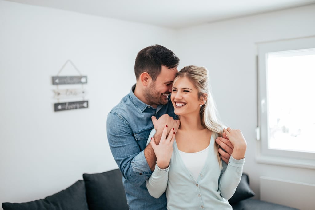 Nuori pari seisoo olohuoneessa lähekkäin. Mies halaa naista takaapäin ja painaa otsansa hänen ohimoaan vasten. Molemmat hymyilevät lämpimästi, ja tilanne huokuu hellyyttä ja onnellisuutta. Taustalla näkyy vaalea sisustus ja ikkuna.