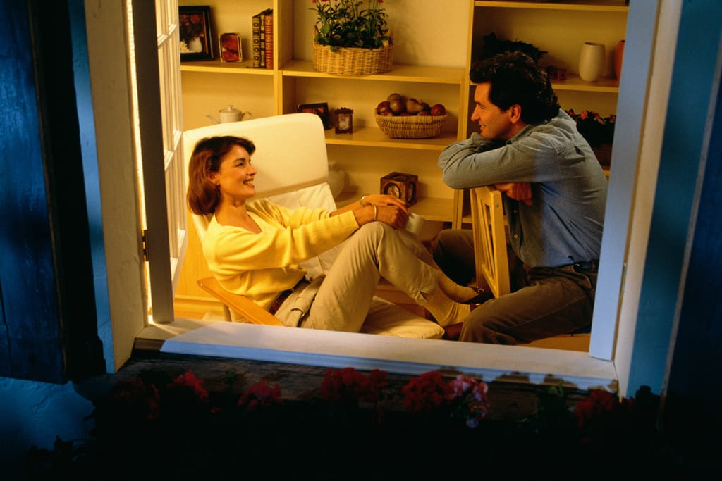 Onnellinen pari istuu vastakkain kodikkaassa olohuoneessa iltahämärässä. He juttelevat ja hymyilevät toisilleen lämpimässä valossa, ympärillä hyllyjä ja kodin yksityiskohtia, jotka korostavat rauhallista ja intiimiä tunnelmaa.