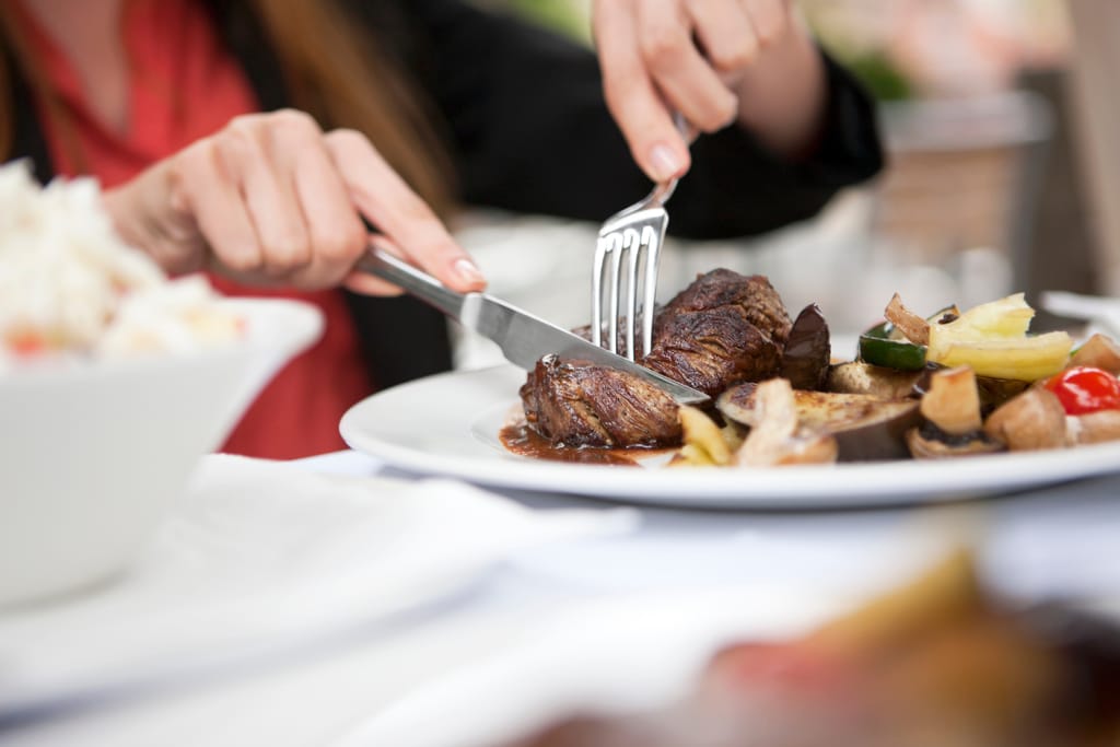 Henkilö leikkaa pihviä lautasella, lisukkeena kasviksia.