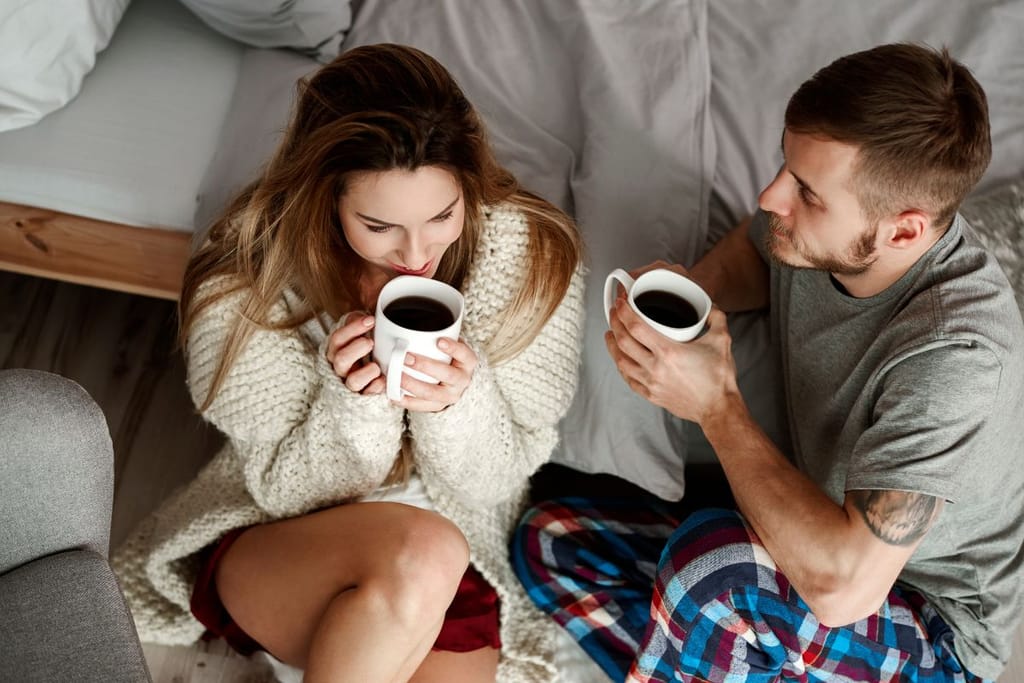 Pariskunta istuu sängyn reunalla ja juo kahvia.
