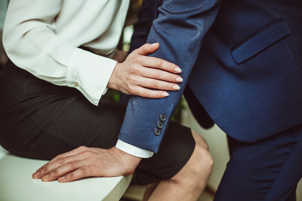 Lähikuva tilanteesta, jossa vaaleaan puseroon pukeutunut henkilö koskettaa tummansiniseen pukuun pukeutuneen henkilön käsivartta; henkilöt istuvat lähekkäin sisätilassa.