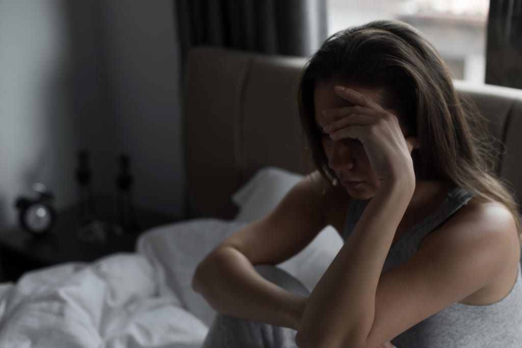 Nainen istuu sängyllä pää käden varassa, katse alaspäin suunnattuna; hänen ilmeensä ja asennonsa viestivät huolta, uupumusta tai emotionaalista ahdistusta hämärässä makuuhuoneessa.