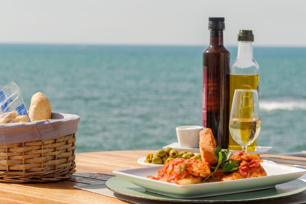 Rantaterassilla katettu pöytä meren äärellä: lautasella tomaattinen kala-annos ja leipää, vieressä oliiviöljy- ja viinipullo, lasi valkoviiniä sekä kori leipää, taustalla avautuu sininen meri.