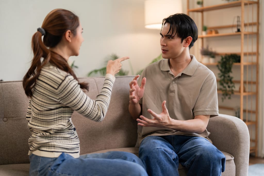 Kaksi aikuista istuu vastakkain sohvalla olohuoneessa ja riitelee. Nainen osoittaa miestä sormella, ja mies vastaa elehtimällä käsillään. Molemmilla on vakava ilme, ja taustalla näkyy kasveja sekä hyllyjä.