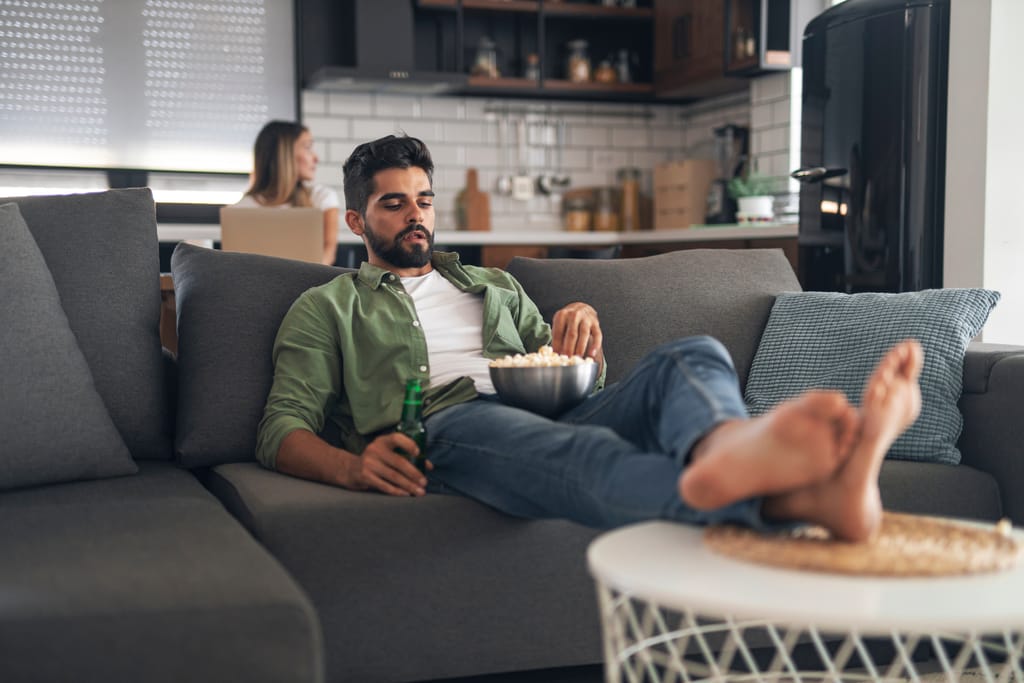 Mies makaa rennosti sohvalla jalat pöydällä, syö popcornia ja pitelee juomapulloa, kun nainen työskentelee kannettavan tietokoneen ääressä keittiössä taustalla.