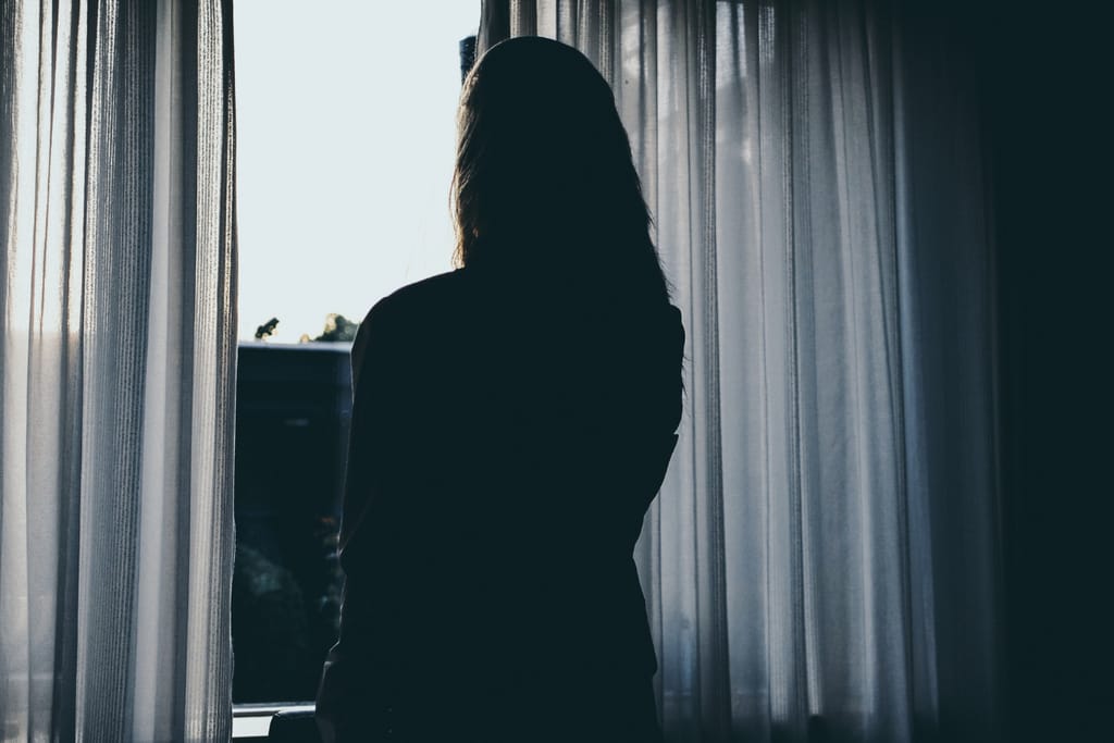 Siluettimainen hahmo seisoo ikkunan edessä puoliksi vedettyjen verhojen välissä ja katsoo ulos kirkkaaseen päivänvaloon. Tunnelma on hiljainen ja etäinen, ja vastavalo korostaa yksinäisyyden ja pohdinnan tunnetta.