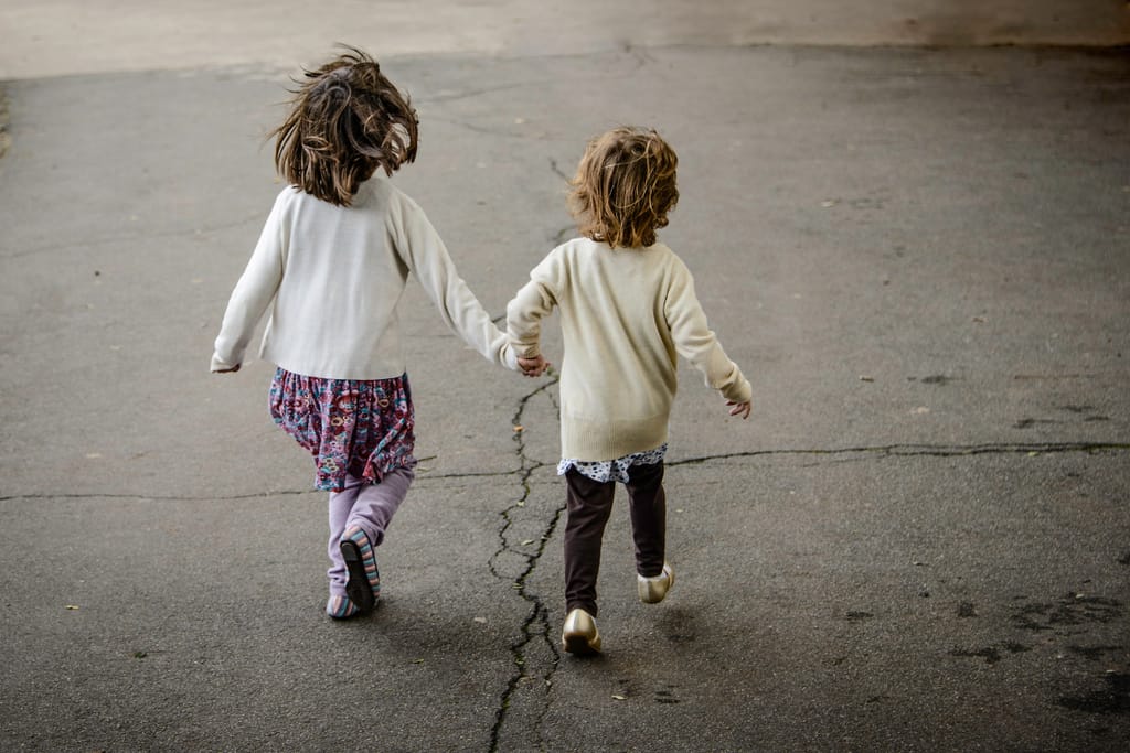 Kaksi pientä lasta kävelee käsi kädessä poispäin kamerasta asfaltilla, toinen hieman toista pidempi. Heillä on vaaleat neuleet ja liike näyttää huolettomalta.