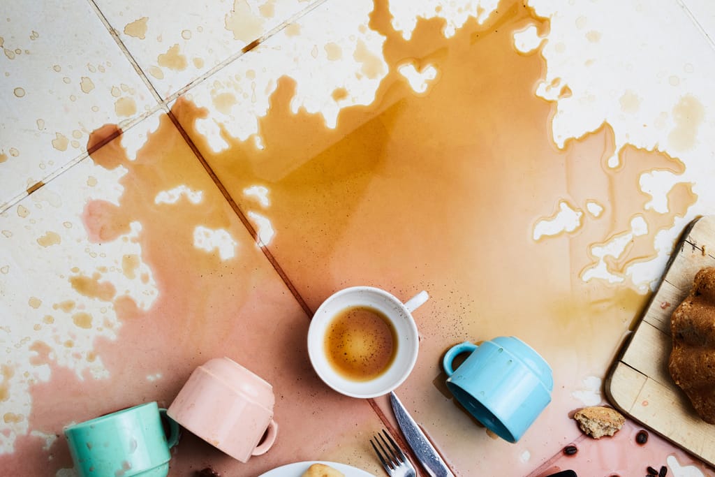 Lattialle ja pöydälle roiskunutta kahvia ja murusia; keskellä valkoinen kahvikuppi, ympärillä kaatuneita värikkäitä mukeja, kahvitahroja ja leivonnaisia.