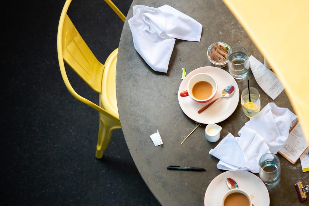 Ylhäältä kuvattu kahvilapöytä, jolla on puoliksi juodut kahvikupit, likaisia lautasia, serviettejä, kuitteja ja vesilaseja; vieressä keltainen tuoli ja sotkuinen kattaus.