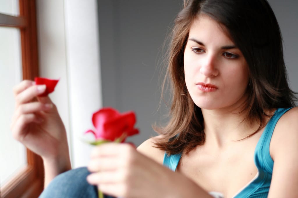 Nainen istuu ikkunan äärellä ja pitelee käsissään punaista ruusua, josta hän on irrottanut terälehden, katse vakavana ja pohtivana. Kuvan tunnelma on surumielinen ja kuvaa rakkauden hiipumista tai suhteen päättymistä symboloivaa hetkeä.