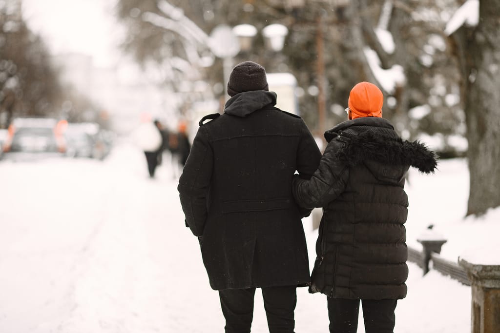 Kaksi aikuista kävelee käsikynkässä lumisella puistokäytävällä selin kameraan. Heillä on paksut talvitakit ja pipot, ja ympärillä näkyy puita sekä katuvaloja. Taustalla kulkee muita ihmisiä, ja talvinen kaupunkiympäristö luo rauhallisen tunnelman.